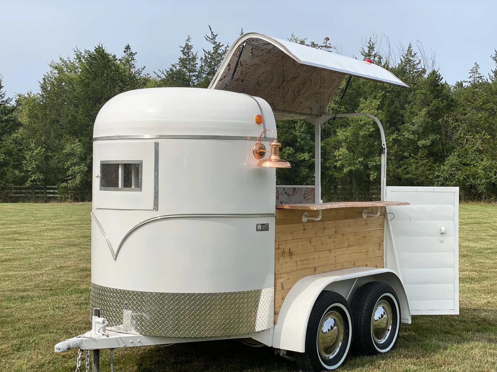 A vintage horse trailer converted into a mobile bar, with cedar counter, copper lamp and a hinged serving hatch